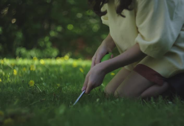 A person wearing a lightcolored top and what appears to be orange shorts is seen kneeling on a vibrant green lawn they are using a small hand tool to interact with the grass possibly weeding or gardening with yellow flowers scattered around them