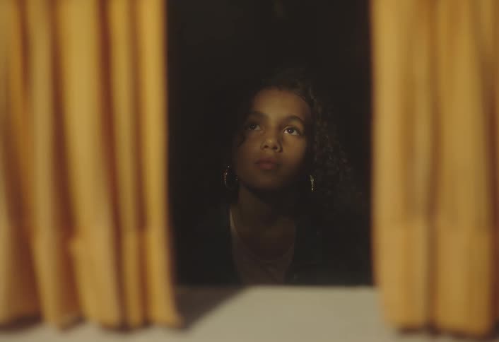 A girl stands behind sheer yellow curtains looking out through a sunlit window