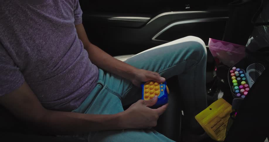 A boy is seen inside a car holding a blue and yellow popit toy which he then puts down he then picks up a yellow bag of snacks and begins to eat from it looking out the car window