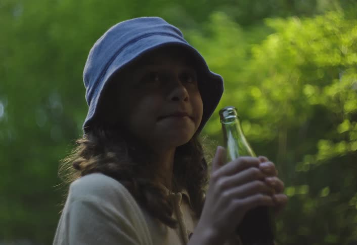 A person wearing a blue bucket hat and a lightcolored top drinking from a green glass bottle with both hands they are outdoors with blurred green foliage in the background suggesting a natural setting