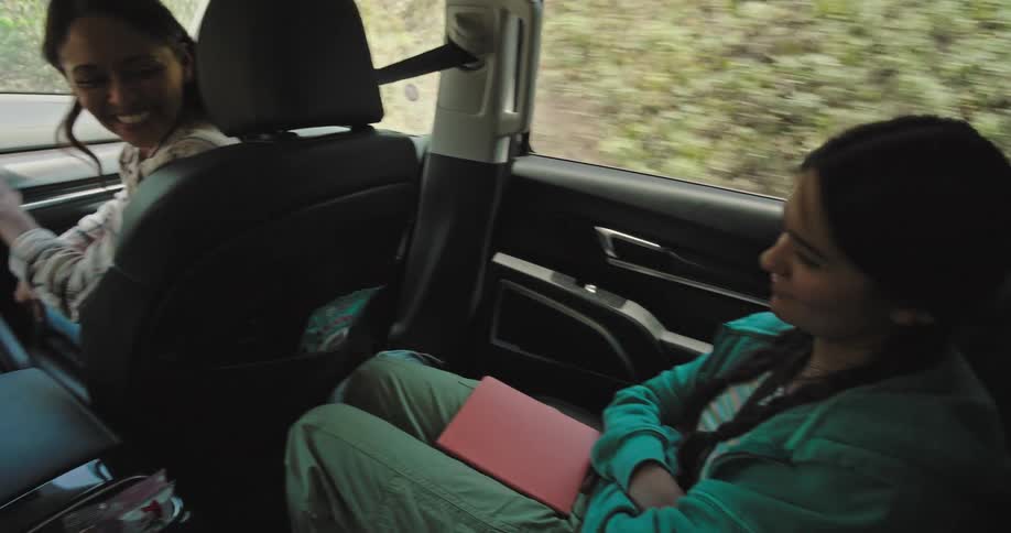 A family is enjoying a car ride with a man driving and a woman in the passenger seat turning to look at the back children are visible in the rear seats and one child peers over the seat looking at the parents in front
