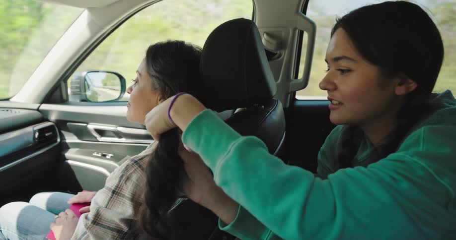 A girl with long dark hair is seen sitting in the back seat of a car expertly braiding the hair of a woman in the passenger seat the woman in the passenger seat has long dark hair and is wearing a plaid shirt