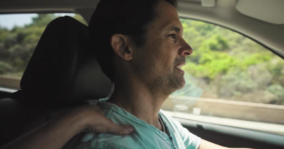 A closeup side profile of a man driving a car is shown with sunlight illuminating his face as he looks forward he has dark hair and a beard and is wearing a light blue shirt