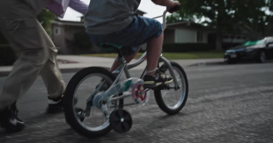 A woman is teaching her son how to ride a bike in the middle of the street with the boy wearing a helmet