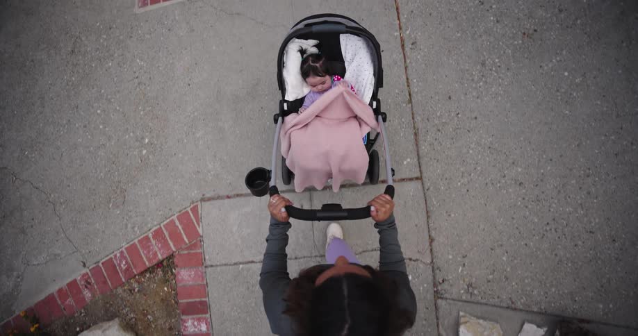 A topdown view shows a person pushing a baby in a stroller the person is wearing a dark top lightcolored pants and white shoes the stroller is black with a pink blanket draped over the baby they are moving along a paved path