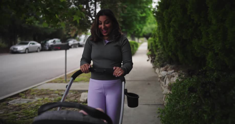 A frontfacing medium shot shows a woman pushing a black stroller down a paved sidewalk the woman is wearing a dark gray longsleeved top and light purple leggings with her hair loose around her shoulders and she is smiling