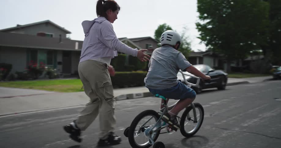 An adult steadies a child riding a bicycle with training wheels on a quiet residential street