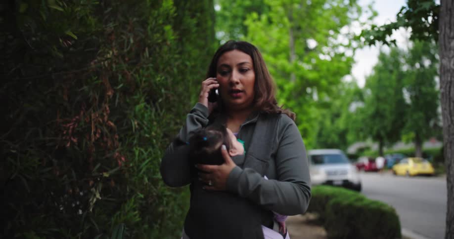 A medium shot captures a woman with long dark hair wearing a grey longsleeved top holding a baby in a black carrier while talking on a phone the babys face is not clearly visible the background consists of lush green foliage