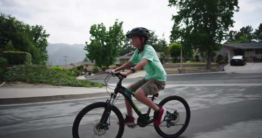 Two people on bicycles one in a blue shirt and the other in a light green shirt are riding on a paved road they navigate past parked cars and houses with one cyclist taking a lead and the other following closely