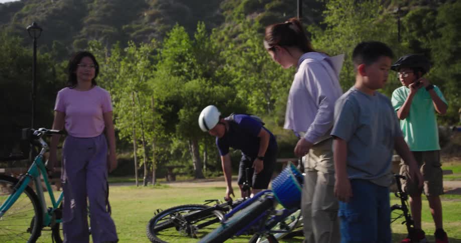 A group of five individuals including two adults and three children are gathered in a park with their bicycles the children are seen putting on and taking off their helmets while some bicycles lie on the grass suggesting they might be taking a break