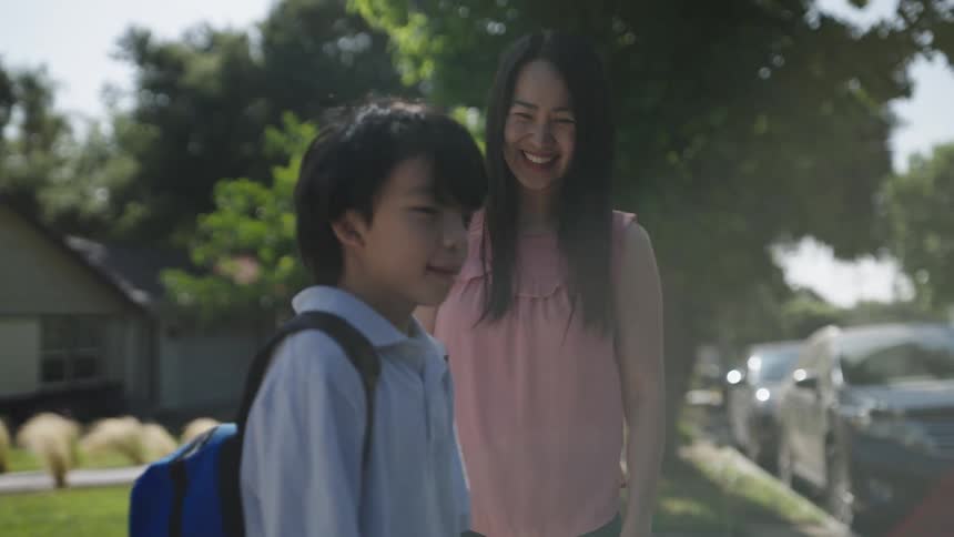 A child with a backpack stands on a sidewalk as his mother wearing a pink sleeveless top and jeans offers encouragement