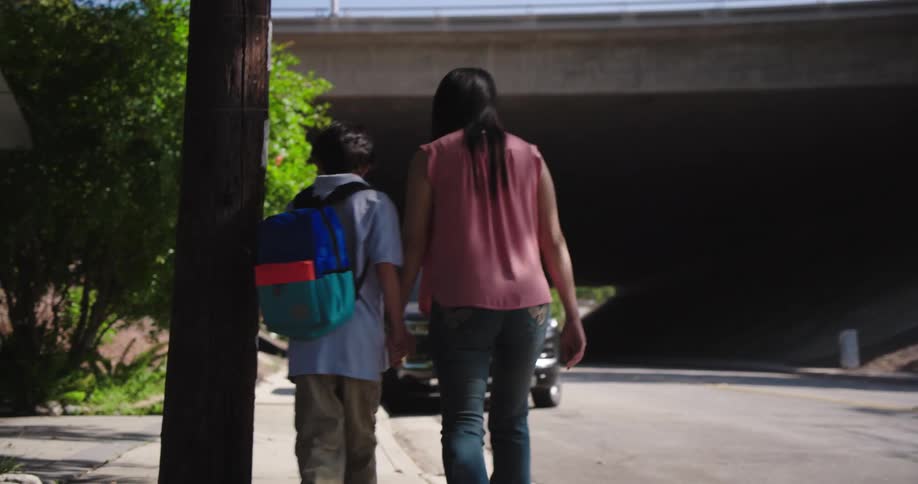 A child wearing a colorful backpack and a woman in a pink shirt and jeans are walking handinhand along a paved path next to a street they are moving away from the camera with trees and bushes lining the side of the path and a dark underpass