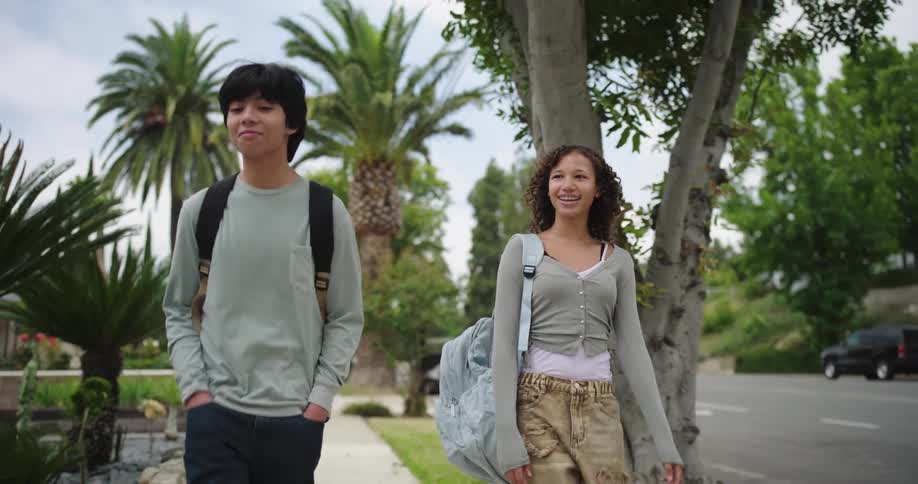 Two teenagers smile and talk while walking along a residential sidewalk on a cloudy day