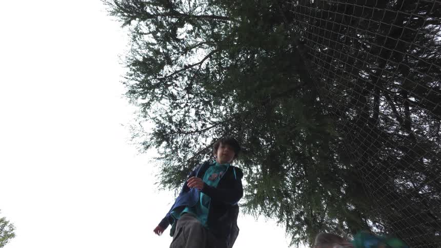 Two young teens take turns jumping while a tree and a chain link fence tower nearby