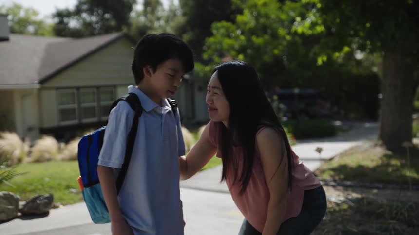 Mother with a happy expression saying goodbye to her son who is walking down the sidewalk with a backpack on his back