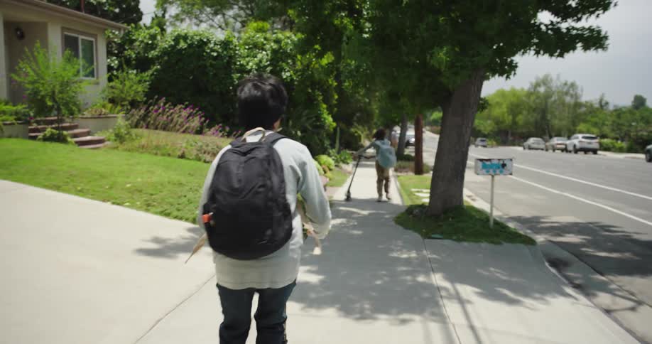 Two children wearing backpacks ride scooters downhill on a sidewalk