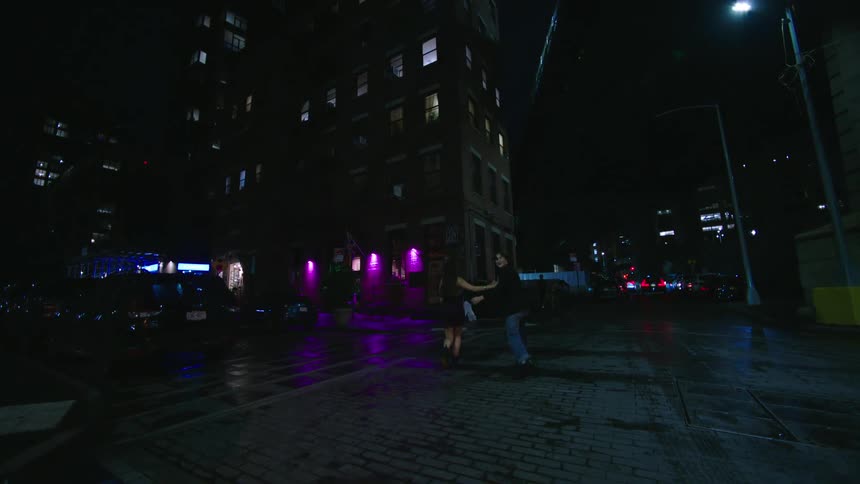A couple walks down a city street at night with wet pavement reflecting colorful lights from buildings