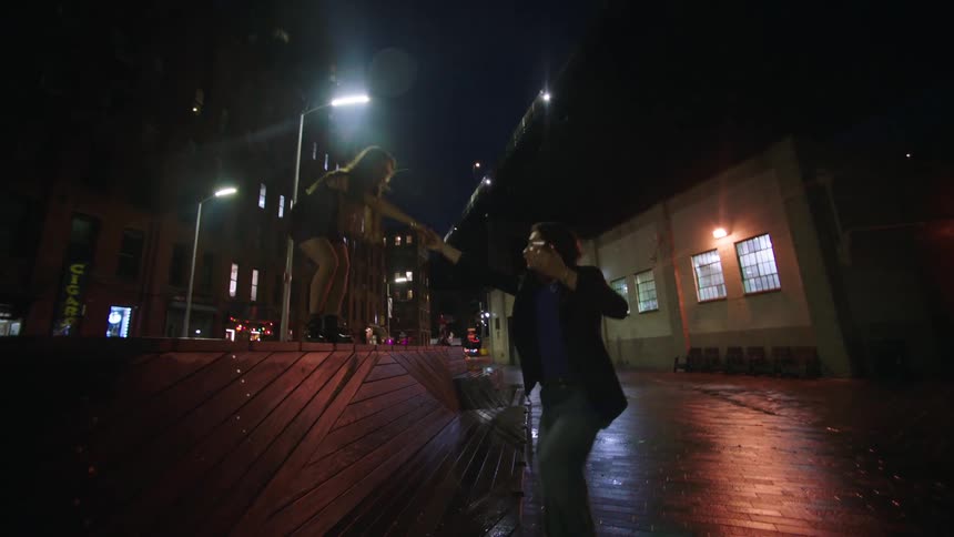 A woman stands on a platform in a city at night and kisses a man while pulling him towards her
