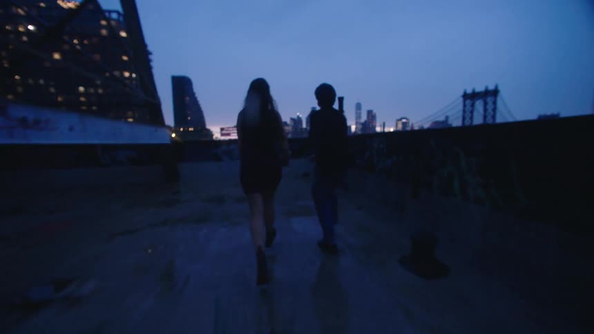 A couple stands on a rooftop at dusk looking out at a city skyline and a bridge