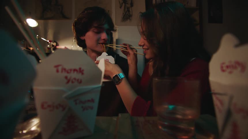 A young couple shares a meal from chinese takeout containers with one feeding the other