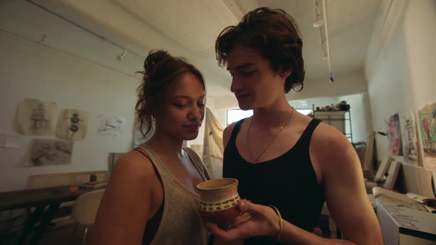 Two young people stand in a welllit room with framed artwork on the walls sharing a tender kiss while holding small cups