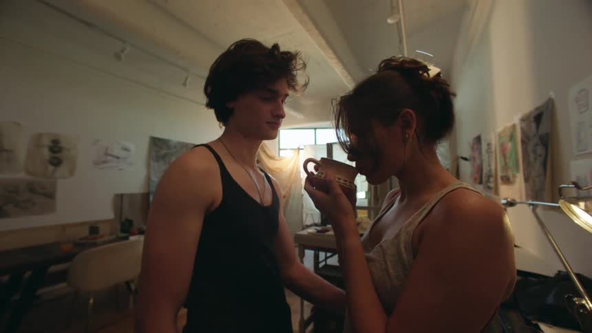 A young couple smiles warmly at each other in an art studio filled with drawings and soft natural light