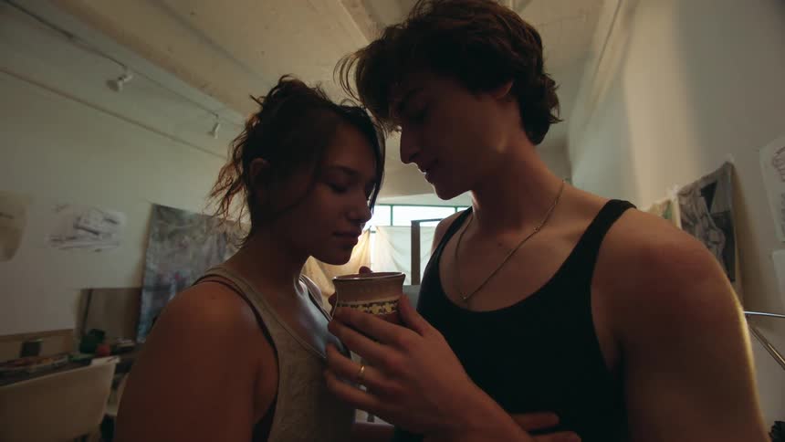 A couple shares a kiss indoors with one holding a cup and the other resting a hand on their partners shoulder