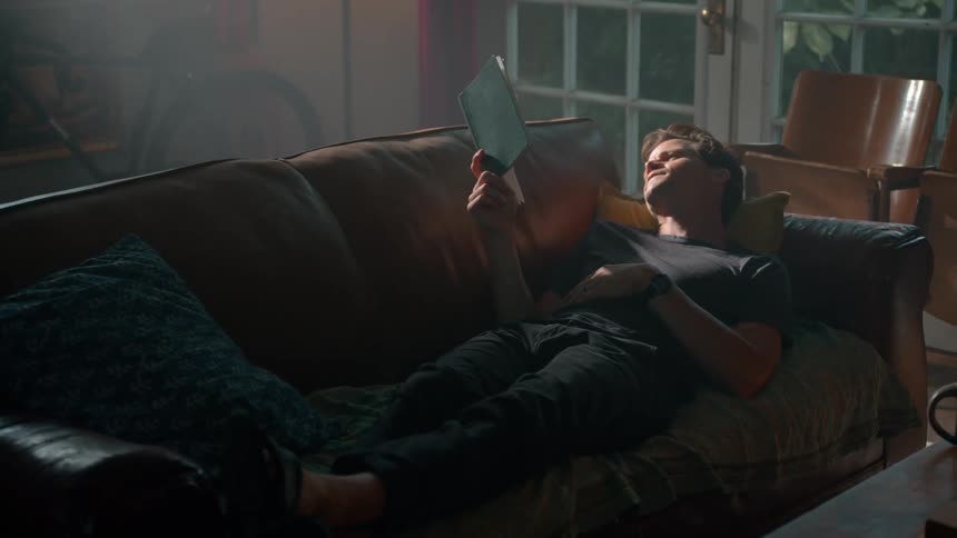A man is reading a book on the couch while laying down
