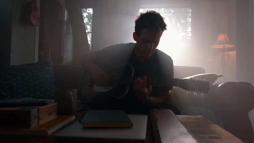 A man sits on a couch strumming a slow melodic tune on his guitar