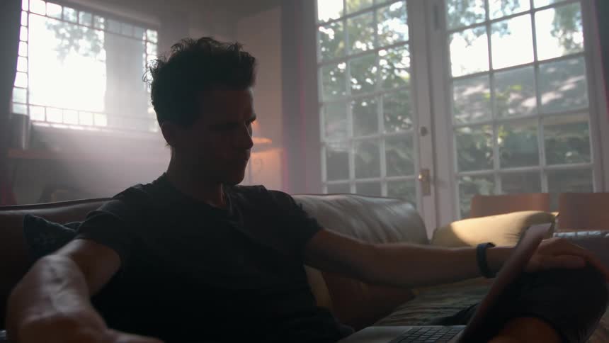A young man uses a laptop while sitting on a couch in a living room during daytime
