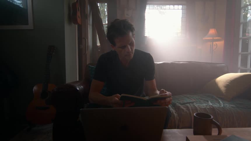 A man flips through a notebook in front of his laptop