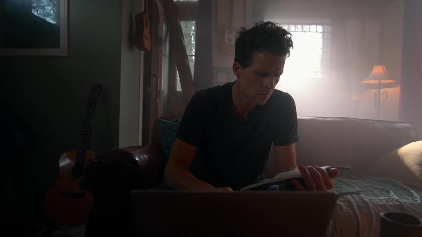 The video shows a man with dark hair wearing a black tshirt sitting on a couch and intently looking at a book in his hands a laptop is on the table in front of him and a lamp provides warm light in the background suggesting a cozy