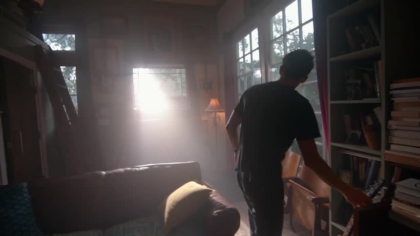 A young man playing a guitar and then placing it on the floor and walking towards a window