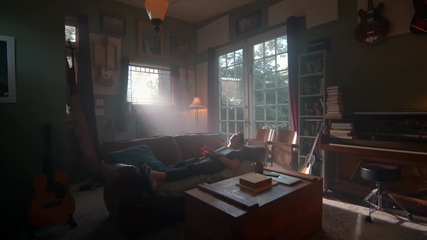 A man lies on a brown leather couch reading a book in a sunlit room with dark green walls surrounded by musical instruments and bookshelves
