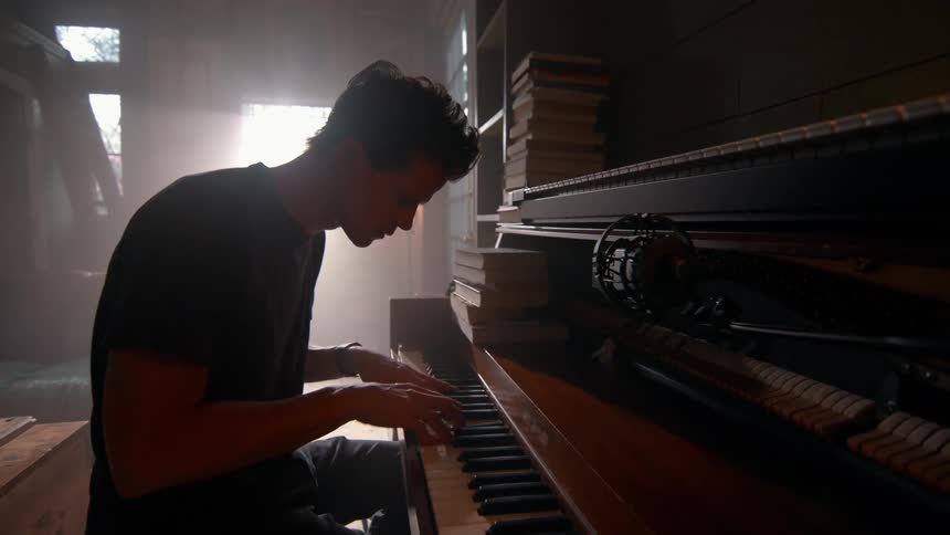 A person plays a piano surrounded by stacked books in a dim room with a guitar hanging on the wall behind
