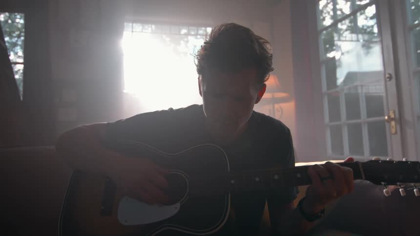 The video shows a man with dark hair wearing a black tshirt sitting on a couch and playing an acoustic guitar sunlight streams in from a large window behind him illuminating the room where other instruments and bookshelves are visible