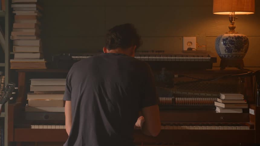 A man plays a piano surrounded by vertical stacks of books with a warm lamp casting soft light from the right
