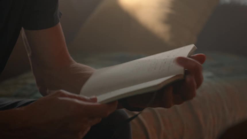 Young man flips through a journal while typing into a laptop computer on a table in a room