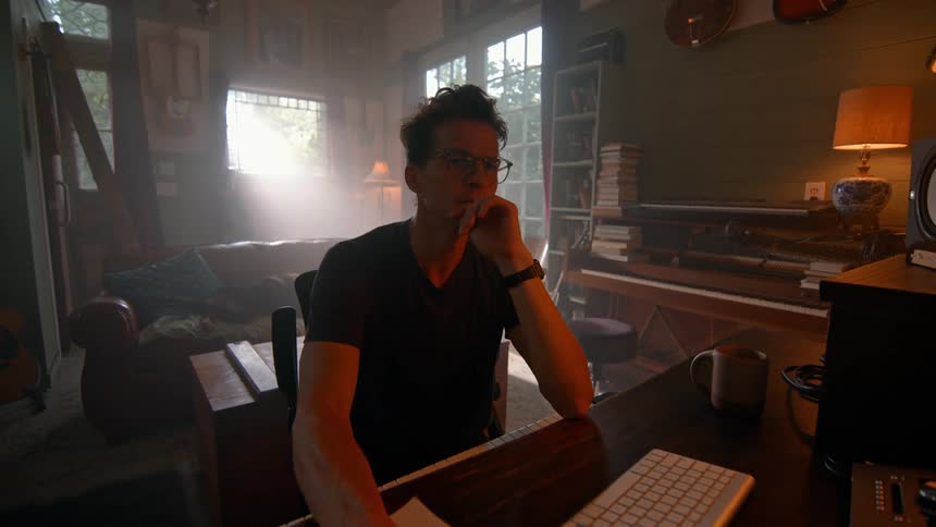 A man sits contemplatively at a table in a living room while using a desktop computer during daytime