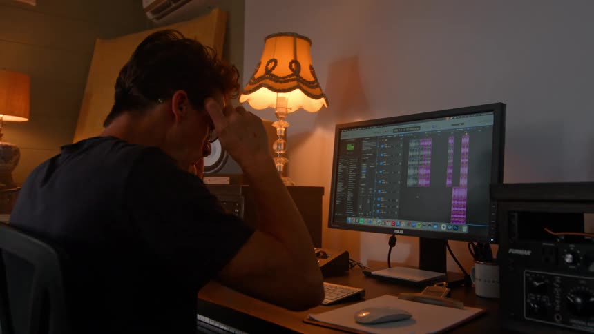 Young male musician sits at a desk to work on a desktop computer in a living room