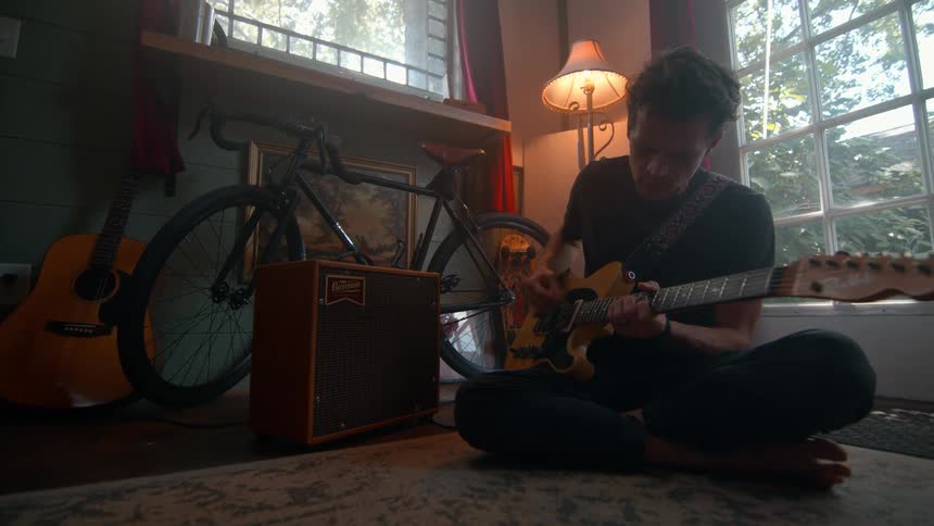A man sitting on the floor plays his orange and black electric guitar