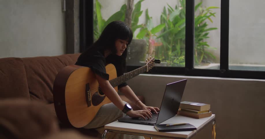 A young woman with long dark hair sits on a brown couch playing an acoustic guitar while looking at a laptop computer on a small white table with a lush plant visible outside a large window