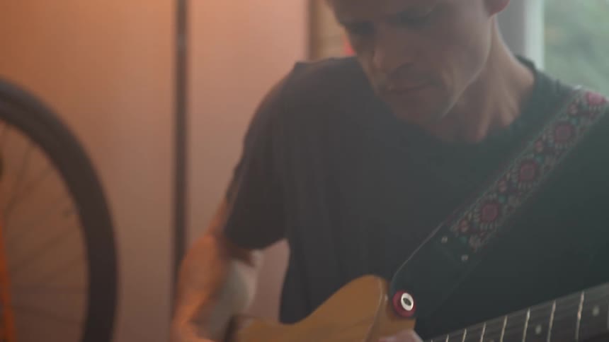 A man plays electric guitar while seated next to a small amp in a cozy room
