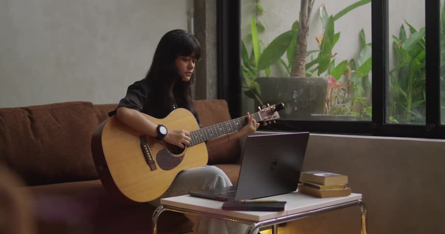 A woman sits on a brown couch playing an acoustic guitar with a laptop and stacked books on a small table near a window that opens to leafy plants outside