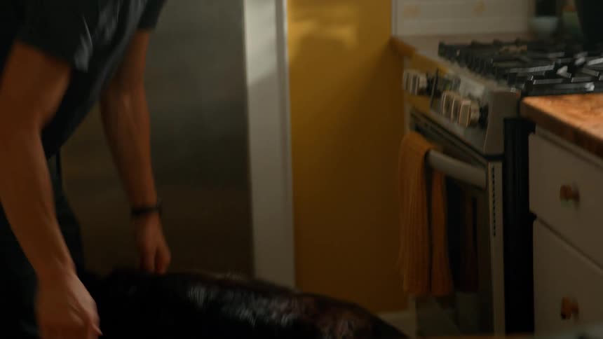 The video shows a man with dark hair wearing a black tshirt standing in a kitchen and interacting with a black dog he bends down to pet and comfort the dog then stands up and reaches for a mug on the counter