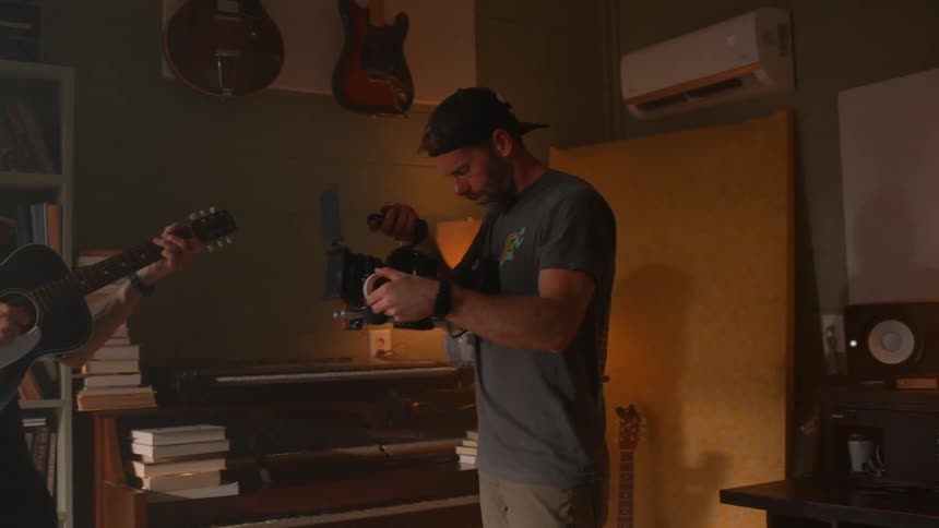 A male videographer records a man playing a guitar in a living room with a camera during daytime