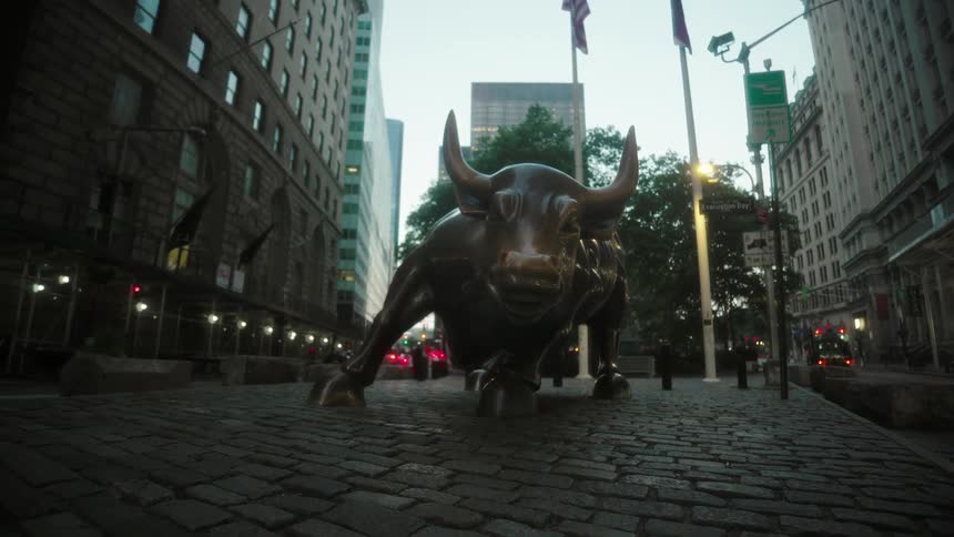 A bronze bull statue stands on a cobblestone street flanked by tall buildings and flags in a dense urban setting