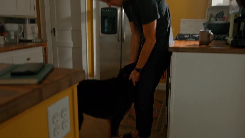 A man kneels in a kitchen hugging a black dog affectionately