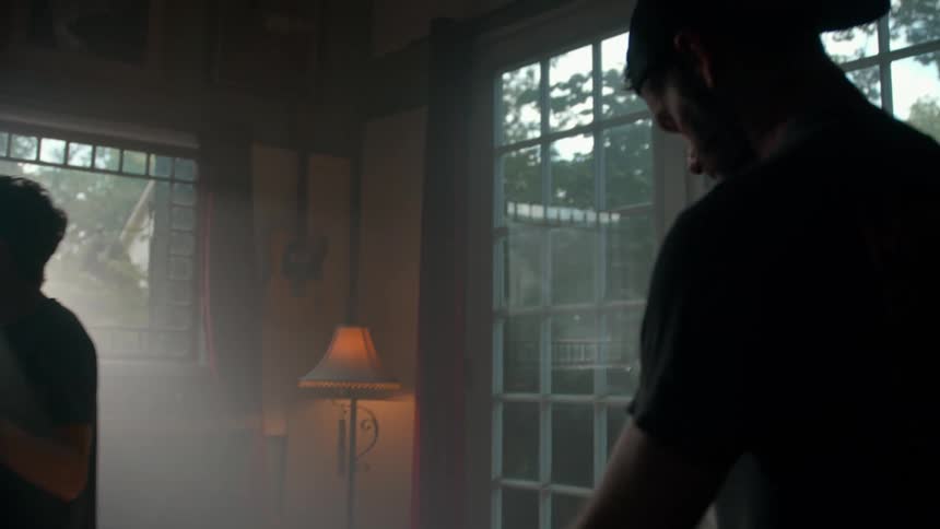 A man is strumming on a guitar while another man films him with a camera in a dimly lit room