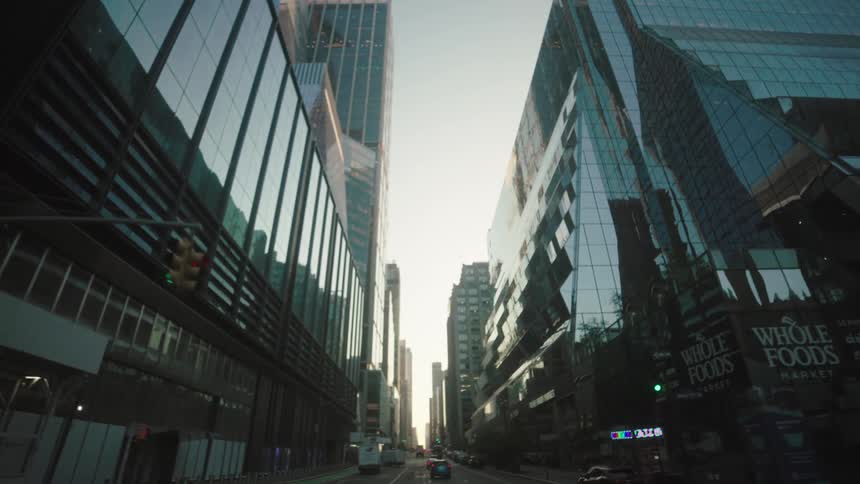 The video filmed from inside a car captures a bustling city street lined with modern highrise buildings and skyscrapers their glass facades reflecting the clear sky as the car moves forward the view shifts from street level to a more upward perspect